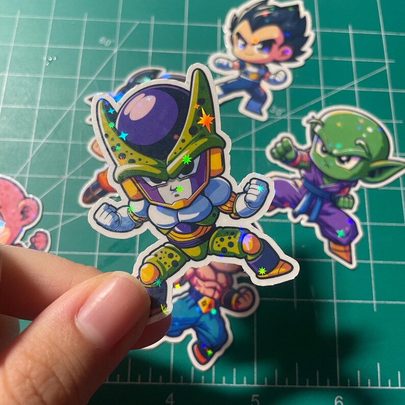 May include: A hand holding a holographic sticker of a cartoon character with a green and purple color scheme. Other stickers of cartoon characters are scattered on a green cutting mat. The stickers have a glittery, iridescent effect.
