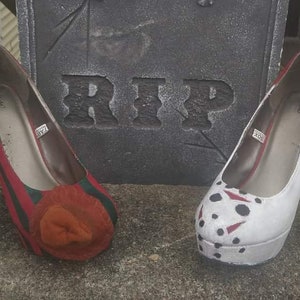 May include: A pair of high-heeled shoes with Halloween-themed designs. One shoe is red and green with a brown flower design, and the other is white with a black and red hockey mask design. The shoes are sitting in front of a tombstone with the word "RIP" on it.