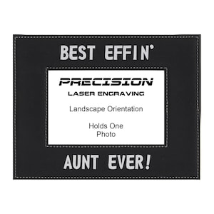 May include: Black leather picture frame with white stitching. The frame has the text "BEST EFFIN' AUNT EVER!" and "PRECISION LASER ENGRAVING" on it. The frame is designed to hold a landscape-oriented photo.
