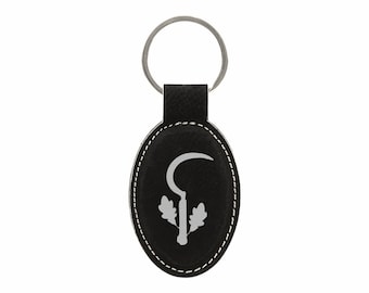 May include: Black oval keychain with a silver key ring. The keychain features a white stitched border and a silver design of a sickle and oak leaves on a black background. The keychain is made of leather.