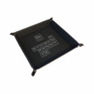 May include: Black leather dice tray with white stitching and a white graphic of a periodic table with the letters "Dm" in the top left corner.