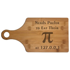 May include: A bamboo cutting board with a handle and a hole for hanging. The board has the text "Nerds Prefer to Eat Their π at 127.0.0.1" etched into the surface. The board is light brown.