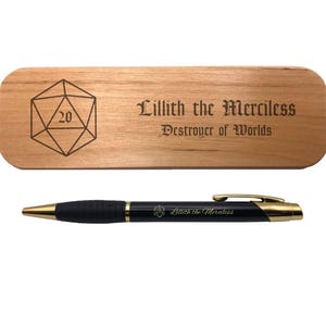 May include: A wooden pen case with the text "Lillith the Merciless Destroyer of Worlds" and a D20 die graphic. A black and gold pen with the same text is below the case.