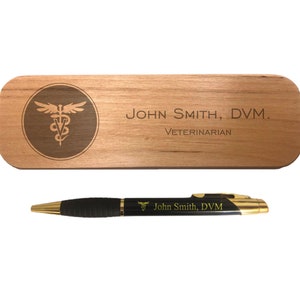 May include: A wooden pen case and pen set. The case is engraved with a medical symbol and the text "JOHN SMITH, DVM. VETERINARIAN." The pen is black with gold accents and also features the name and medical symbol.