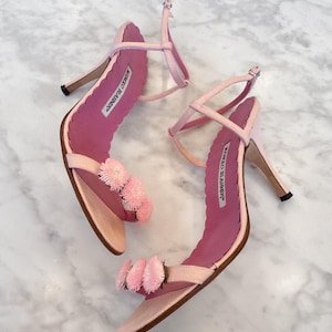 Puede incluir: Sandalias de tacón alto rosa claro con un diseño de tiras delicadas. Las sandalias presentan un detalle decorativo de pompón en la tira del dedo y un borde festoneado. El nombre de la marca "MANOLO BLAHNIK" es visible en la plantilla.