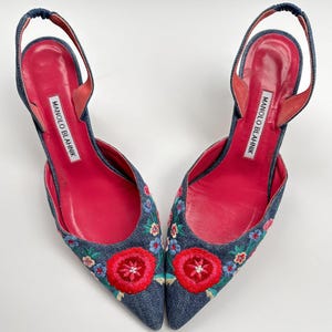 May include: A pair of denim slingback heels with floral embroidery. The shoes feature a pointed toe, a red leather insole, and a strap around the ankle. The heels have the brand name "MANOLO BLAHNIK" printed on the insole.