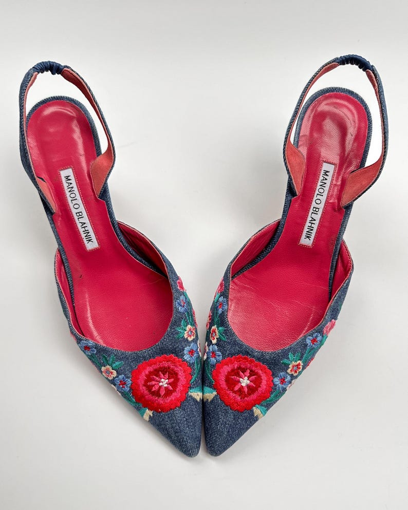 May include: A pair of denim slingback heels with floral embroidery. The shoes feature a pointed toe, a red leather insole, and a strap around the ankle. The heels have the brand name "MANOLO BLAHNIK" printed on the insole.