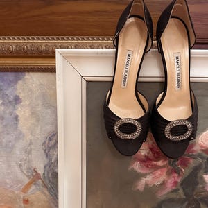Puede incluir: Un par de zapatos de tacón alto de satén negro con laterales abiertos y una hebilla decorativa de cristal. Los zapatos tienen el nombre de la marca "Manolo Blahnik" impreso en la plantilla.