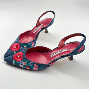 May include: A pair of denim slingback heels with floral embroidery. The shoes feature a pointed toe, a small heel, and a pink insole. The floral embroidery includes red, pink, blue, and green colors.