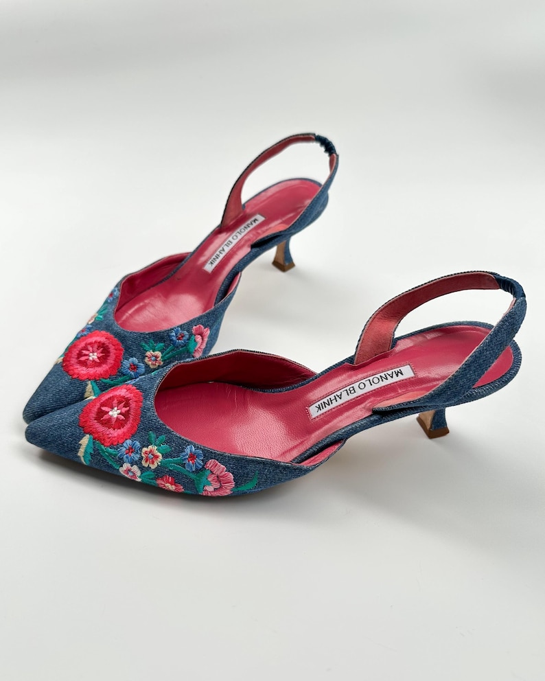 May include: A pair of denim slingback heels with floral embroidery. The shoes feature a pointed toe, a small heel, and a pink insole. The floral embroidery includes red, pink, blue, and green colors.
