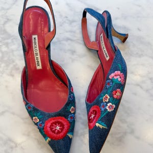 May include: A pair of denim slingback heels with floral embroidery. The shoes feature a pointed toe, a thin strap, and a small heel. The interior is a bright pink color, and the brand name "MANOLO BLAHNIK" is visible.