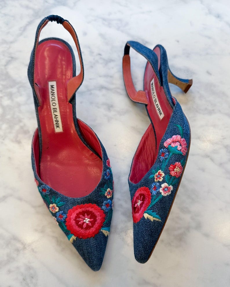 May include: A pair of denim slingback heels with floral embroidery. The shoes feature a pointed toe, a thin strap, and a small heel. The interior is a bright pink color, and the brand name "MANOLO BLAHNIK" is visible.