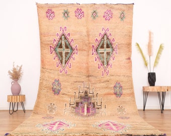 May include: A vintage Moroccan rug with a beige background and intricate geometric patterns in shades of pink, purple, blue, and brown. The rug has a distressed look and frayed edges.