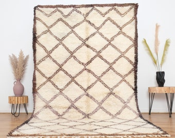 May include: A large, cream-colored rug with a geometric diamond pattern in shades of brown. The rug has a thick, textured pile and a fringed edge. The design features a repeating pattern of intersecting lines, creating a grid-like appearance. The rug is displayed against a white wall.