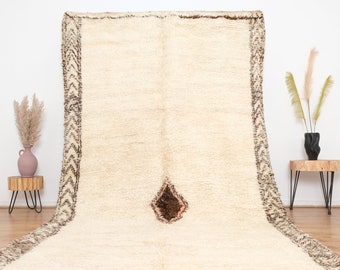 May include: A white Moroccan rug with a brown and white geometric border and a brown and orange diamond shape in the center. The rug has a fringe border.