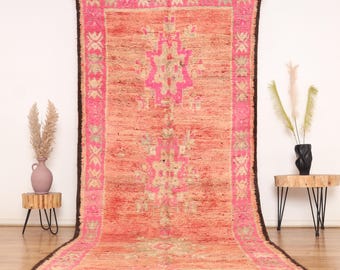 May include: A rectangular, hand-woven rug with a central geometric design in shades of pink and beige. The rug has a dark brown border and fringe, and is displayed on a wooden floor. The rug's dimensions are not visible.
