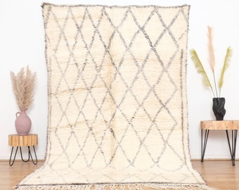 May include: A white Moroccan rug with a diamond pattern in gray. The rug has a fringe border and is laid out on a wooden floor.