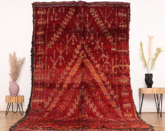 May include: A large, rectangular, hand-woven rug in shades of red, orange, and brown. The rug features a geometric pattern with diamond and cross-like shapes. The edges are finished with a dark fringe.