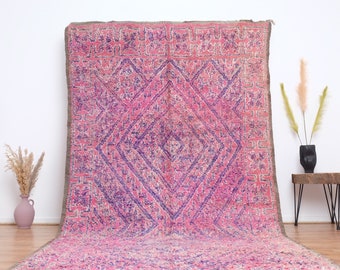 May include: A pink and purple Moroccan rug with a geometric pattern. The rug has a diamond shape in the center with a smaller diamond shape inside. The rug is made of wool and has a fringe on the edges.