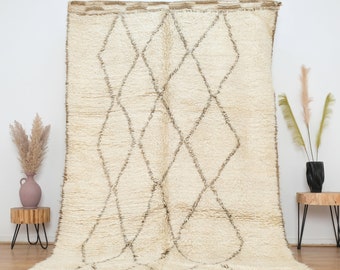 May include: Cream-colored, textured rug with a geometric diamond pattern in brown. The rug has a fringe along the bottom edge and is displayed against a white wall. Decorative elements include vases and small wooden tables.