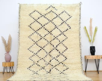 May include: A white Moroccan rug with a black diamond pattern. The rug has a fringe border and is made of wool.