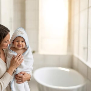 Könnte beinhalten: Ein weißes Baby-Kapuzenhandtuch mit hellblauer Einfassung und gesticktem Design. Das Handtuch ist um ein lächelndes Baby gewickelt. Auf der Kapuze steht "Petit Joueur".