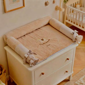 May include: A beige baby changing pad with a rocking horse embroidery and the text "NON BÉBÉ". The pad has a soft, plush surface and is surrounded by padded bolsters. The bolsters are accented with a checkered fabric and bows. The changing pad is placed on a white dresser.