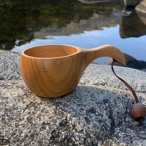 May include: A wooden Kuksa cup with a curved handle and a leather cord with a wooden bead. The cup is made of light brown wood and sits on a grey rock. The background shows water and rocks.