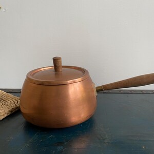 May include: A copper saucepan with a wooden handle and lid. The pot has a rounded shape and a small wooden knob on the lid. The handle is long and angled. The pot is sitting on a dark blue surface.