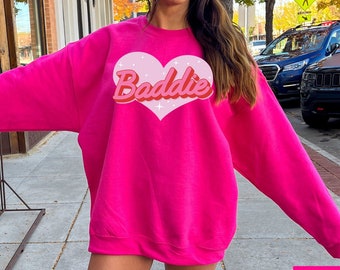 May include: A pink sweatshirt with the word "Baddie" in a retro font inside a pink heart. The sweatshirt is being worn by a person with white sneakers.