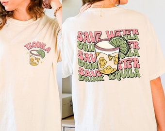 May include: Ivory-colored t-shirt with a tequila-themed graphic. The front features a cocktail illustration with the word "TEQUILA". The back displays the phrase "SAVE WATER DRINK TEQUILA" in a repeating pattern, with a cocktail illustration.