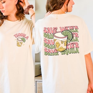 May include: Ivory-colored t-shirt with a tequila-themed graphic. The front features a cocktail illustration with the word "TEQUILA". The back displays the phrase "SAVE WATER DRINK TEQUILA" in a repeating pattern, with a cocktail illustration.