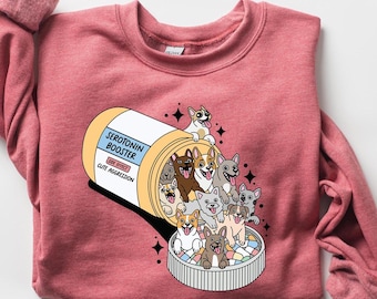 May include: A dusty rose sweatshirt featuring a graphic of a pill bottle labeled "Serotonin Booster" with various dog breeds spilling out. The design includes the text "Cute Aggression".