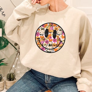 May include: A light beige sweatshirt with a large smiley face graphic. The smiley face is filled with small images of ghosts, pumpkins, and hearts. The text "FREE GIFT WRAP" is in a red circle with a red bow on top. The text "Sand" is in a white rectangle below the circle.
