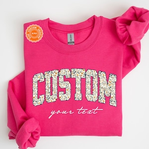 May include: Hot pink sweatshirt with the word "CUSTOM" in a floral design. The text "your text" is below the word "CUSTOM". The sweatshirt has the brand name "GILDAN" on it.