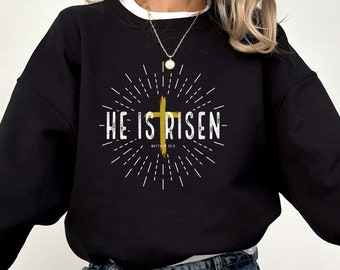 May include: Black sweatshirt with a white graphic design featuring a gold cross and the text "He is Risen" with the scripture reference "Matthew 28:6".