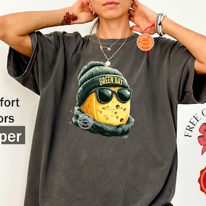 May include: Dark grey t-shirt featuring a cartoon cheese character wearing a Green Bay beanie and sunglasses. The shirt has the words "Comfort Colors" and "Pepper" printed on the left side. A gift wrap and color availability badge are also visible.
