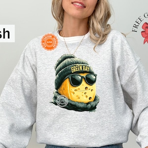 May include: Ash gray sweatshirt featuring a graphic of a cheese wedge wearing a green beanie with "GREEN BAY" text, sunglasses, and a scarf. The sweatshirt has a crew neck and long sleeves. The words "FREE GIFT WRAP" are printed on the upper right.