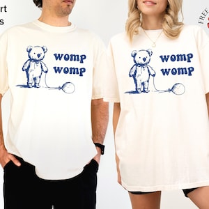 May include: Two ivory t-shirts with a cartoon bear holding a balloon and the text "womp womp" printed on the front.