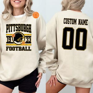 May include: Sand-colored sweatshirt with "PITTSBURGH 1933 FOOTBALL" text and a football helmet graphic. The back features "CUSTOM NAME" and the number "00" in black with gold trim. Gildan brand.