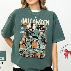 May include: A teal blue t-shirt with a vintage-style graphic of characters from the Halloween movie franchise. The graphic includes the text "Halloween", "The Night Come Home!" and "John Myers".