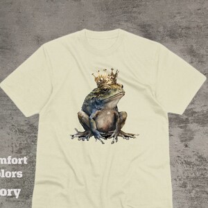 May include: A beige t-shirt with a graphic of a frog wearing a gold crown. The frog is sitting on its hind legs with its front legs crossed. The frog is green and brown with a white belly.