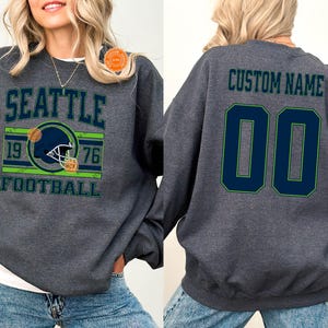 May include: Dark heather gray sweatshirt with "SEATTLE FOOTBALL" text and a football helmet graphic. The back features "CUSTOM NAME" and the number "00" in navy blue with green trim. A unisex retail size garment.