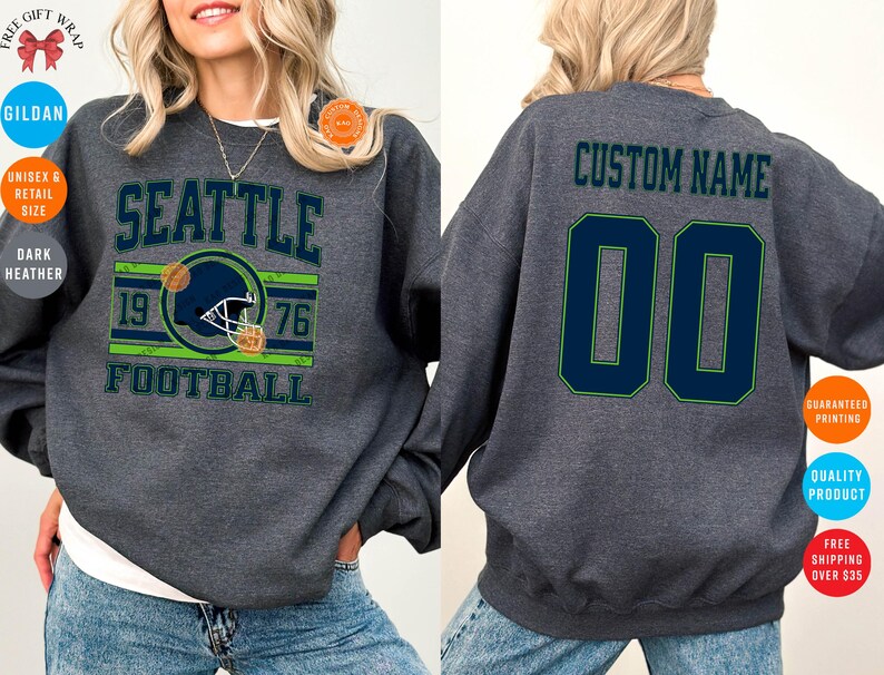 May include: Dark heather gray sweatshirt with "SEATTLE FOOTBALL" text and a football helmet graphic. The back features "CUSTOM NAME" and the number "00" in navy blue with green trim. A unisex retail size garment.