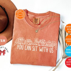 May include: A coral-colored t-shirt with the phrase "YOU CAN SIT WITH US" in white. The shirt features a crew neck and a graphic of people sitting. A gold necklace is draped on the shirt.