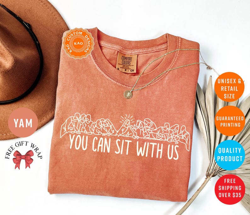May include: A coral-colored t-shirt with the phrase "YOU CAN SIT WITH US" in white. The shirt features a crew neck and a graphic of people sitting. A gold necklace is draped on the shirt.