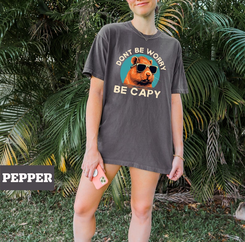 May include: A dark gray t-shirt with a graphic of a capybara wearing sunglasses. The graphic is in a circle with the text "Don't be worry be capy" in a vintage font.