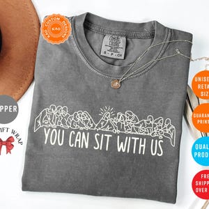 May include: A gray Comfort Colors t-shirt with the text "YOU CAN SIT WITH US" in white. The shirt features a line drawing of people sitting together. A gold necklace is draped across the shirt.