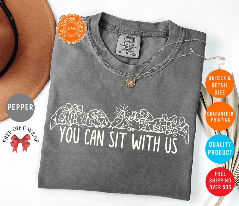May include: A gray Comfort Colors t-shirt with the text "YOU CAN SIT WITH US" in white. The shirt features a line drawing of people sitting together. A gold necklace is draped across the shirt.