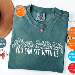 May include: Blue spruce Comfort Colors t-shirt with the text "YOU CAN SIT WITH US" in white. The shirt features a white graphic of people sitting. A gold necklace is draped on the shirt. The shirt is a unisex retail size.